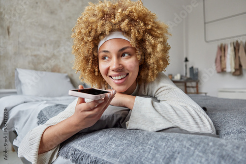 Pleased relaxed curly haired woman rests in bedroom on bed enjoys modern technologies being at home during weekend smiles gladfully uses virtual assistant on mobile phone records voice message