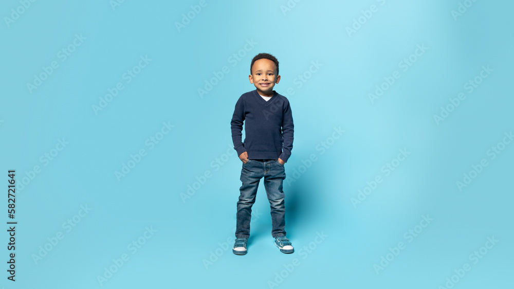 Naklejka premium Happy childhood. Full length shot of black little boy holding hands in pockets, blue background, panorama, free space