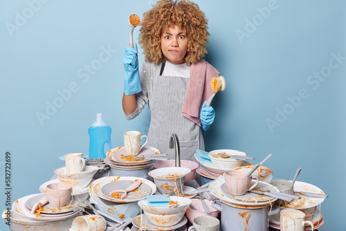 Obraz na plátně Irritated curly haired housewife holds brush for washing uo looks angrily has much work to do wears striped apron and rubber gloves stands near sink full of dirty dishes isolated over blue background