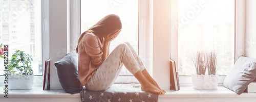 Canvas Print Depressed woman covering face with hands and crying while sitting on the window