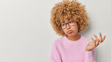 Confused hesitant woman shrugs shoulders feels unaware listens something in displeasure purses lips wears spectacles and pink knitted jumper isolated over white background blank space aside for text