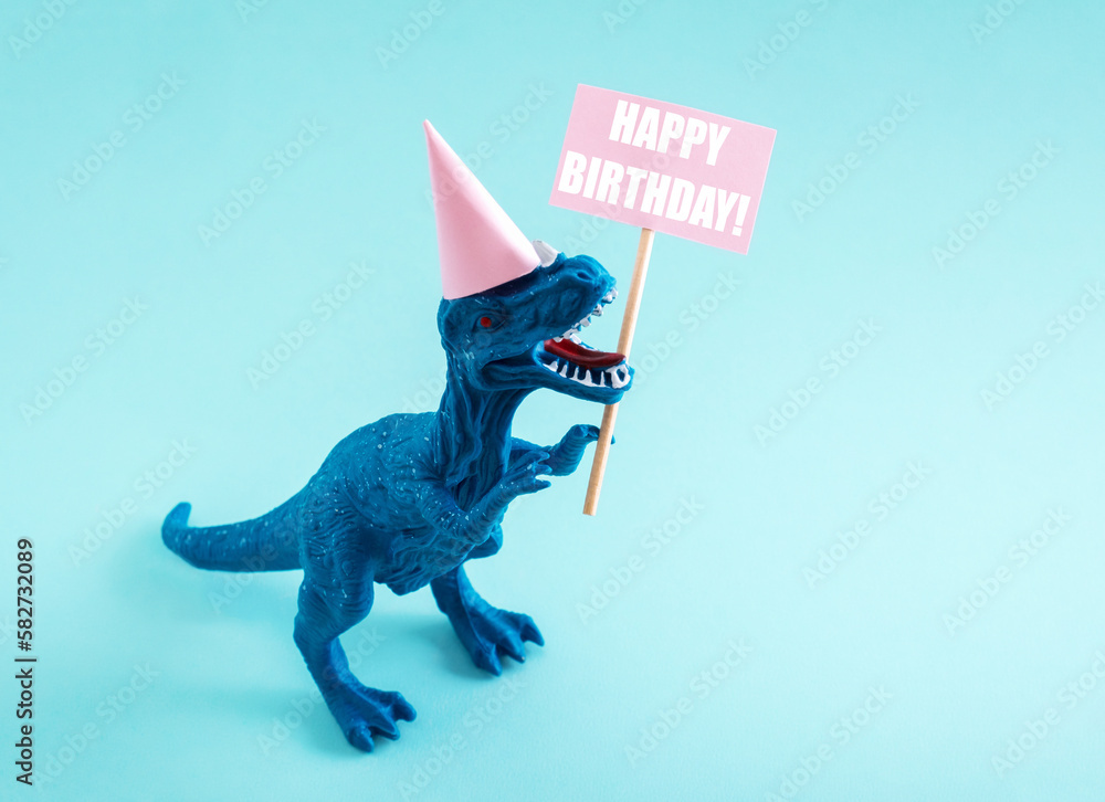 Fototapeta premium Cute dinosaur in birthday hat holding Happy Birthday sign on blue background. Cute birthday greeting card idea concept.