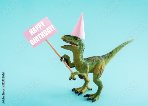 Cute dinosaur in birthday hat holding Happy Birthday sign on blue background. CUte birthday greeting card idea concept. © dvulikaia