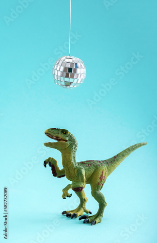 Happy cute green toy dinosaur dancing under disco ball on blue background. Minimal creative art poster. © dvulikaia