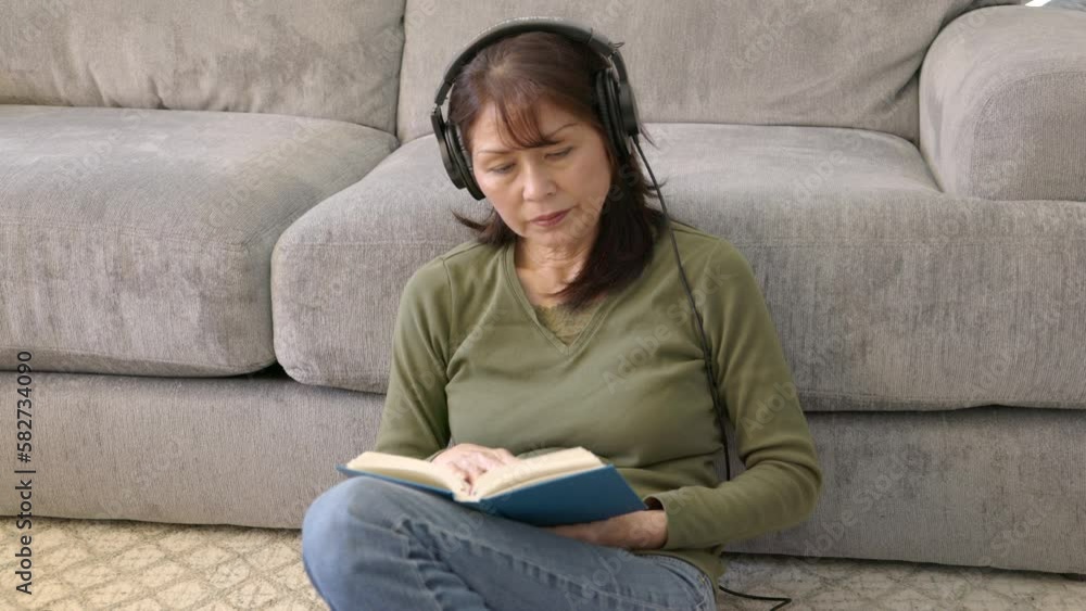 Mature woman wearing headphones sitting on floor resting against sofa reading book Mature woman wearing headphones sitting on floor resting against sofa reading book