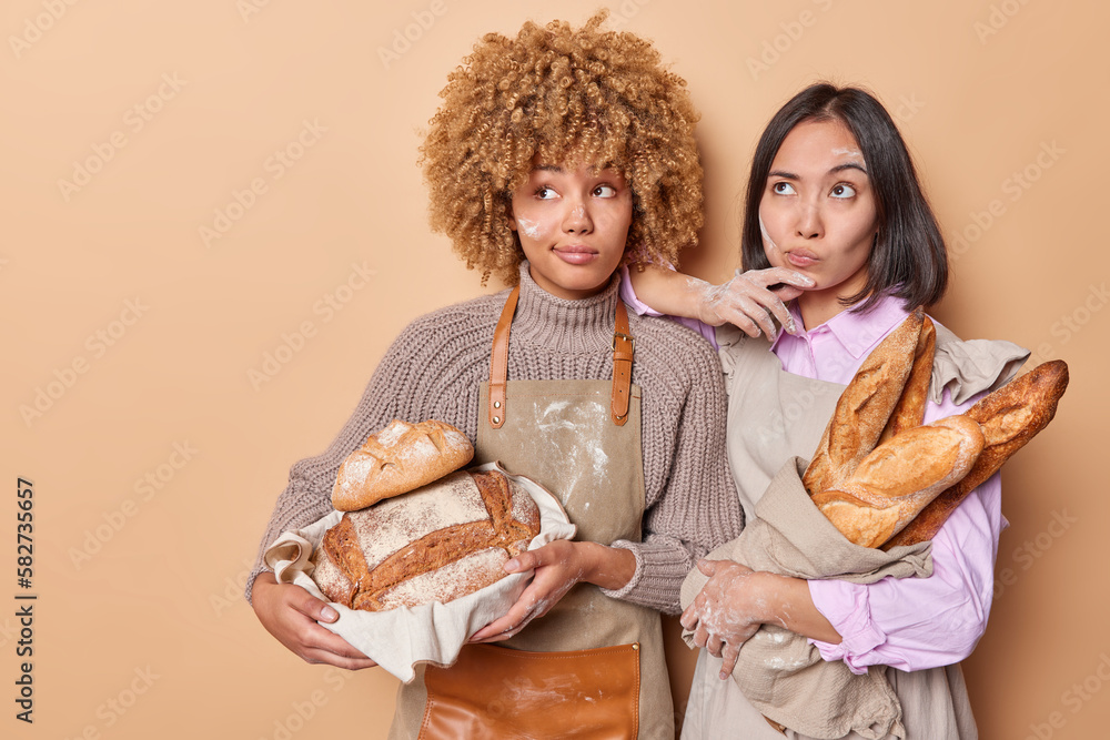 Pensive female bakers work in bakery shop holds assortment of freshly baked bread focused aside stand closely to each other isolated over brown background. Fresh fragrant sourdough delicious bread Pensive female bakers work in bakery shop holds assortment of freshly baked bread focused aside stand closely to each other isolated over brown background. Fresh fragrant sourdough delicious bread