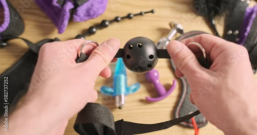 Male hands inspect a bdsm ball gag for erotic games. POV view, a bunch of toys from the sex shop are on the table.