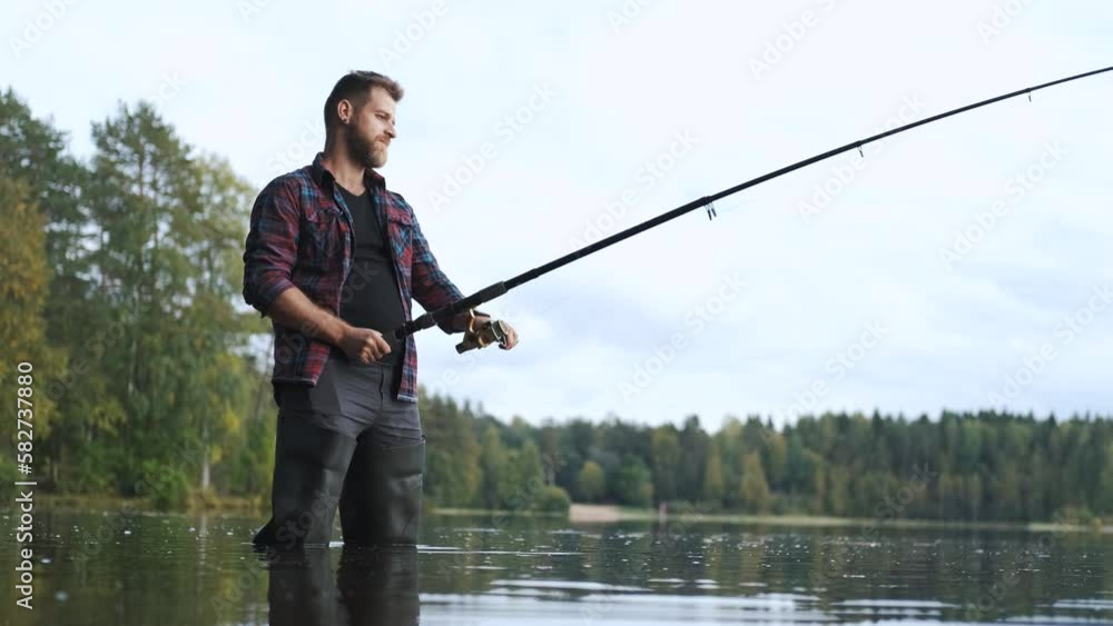 a fisherman in high boots stands in the water of the lake with a fishing rod and hooks a fish a fisherman in high boots stands in the water of the lake with a fishing rod and hooks a fish
