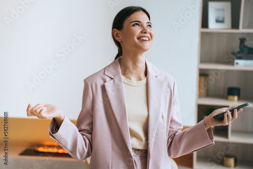Excited Asian young businesswoman in suit  laughing holds phone toothy smiles looks up grateful for success thanks God. Happy brunette girl enjoying wealthy life at home against fireplace, bookshelf,