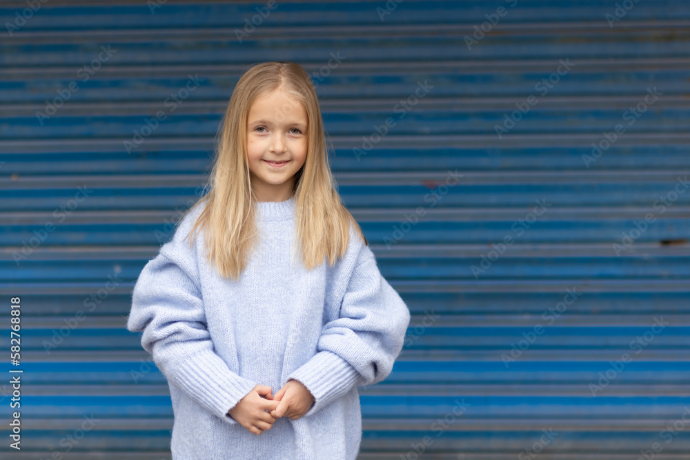 Cute little caucasian girl seven years old with blonde hair on blue background. Kid wearing stylish cozy knitted sweater. Happy Child smiling