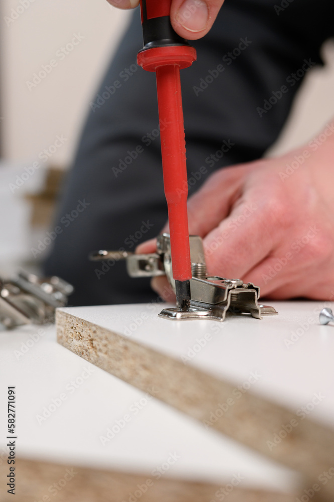 Furniture assembly. A worker is screwing a hinge into a wooden cabinet door with a screwdriver, close-up. Adjustment of fittings, door hinges. The concept of installing furniture, home renovation.