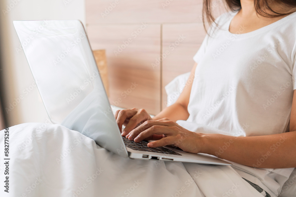 Closeup of woman sitting on bed at home while working on computer. Technology and lifestyle, relaxing at home, work from home concept. Copy space