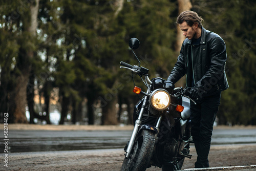 male motorcyclist in a leather motorcycle jacket stands by a motorcycle on a forest road. Motorcycle cafe racer