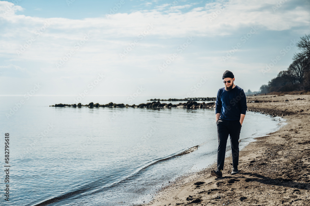 cooler junger Mann mit Bart, Sonnenbrille und Mütze geht bei blauem Himmel am Strand entlang und denkt nach, Blick nach unten. cooler junger Mann mit Bart, Sonnenbrille und Mütze geht bei blauem Himmel am Strand entlang und denkt nach, Blick nach unten.