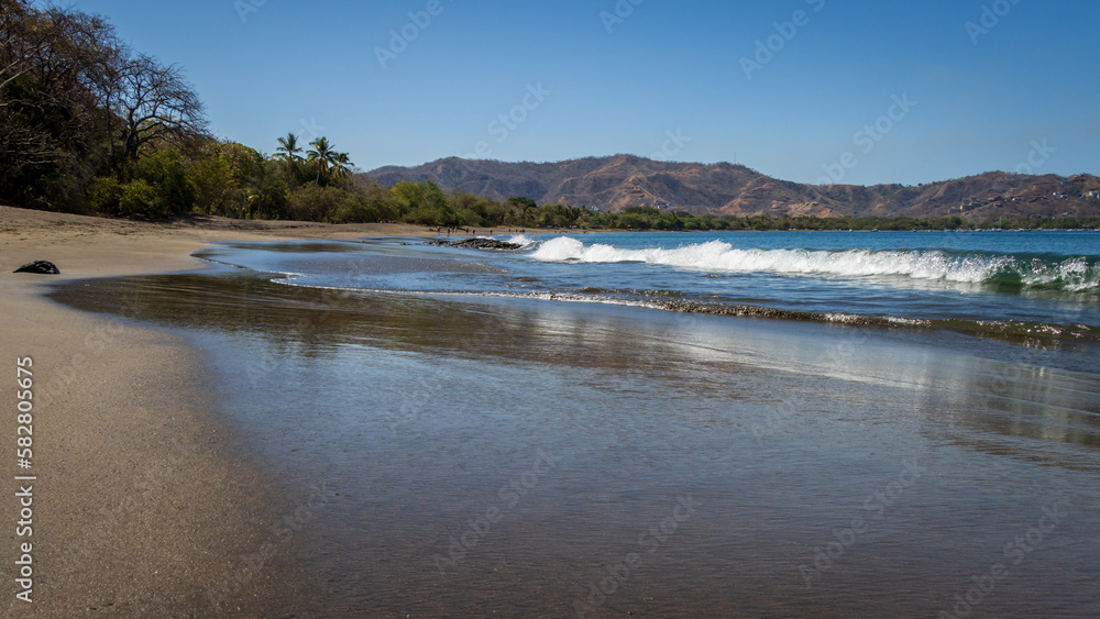 A peaceful tropical image of waves crashing onto a smooth sand beach in Costa Rica. In the distance are hills and palm trees. The beach is mainly deserted of people. 