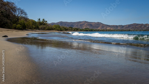 A peaceful tropical image of waves crashing onto a smooth sand beach in Costa Rica. In the distance are hills and palm trees. The beach is mainly deserted of people. 
