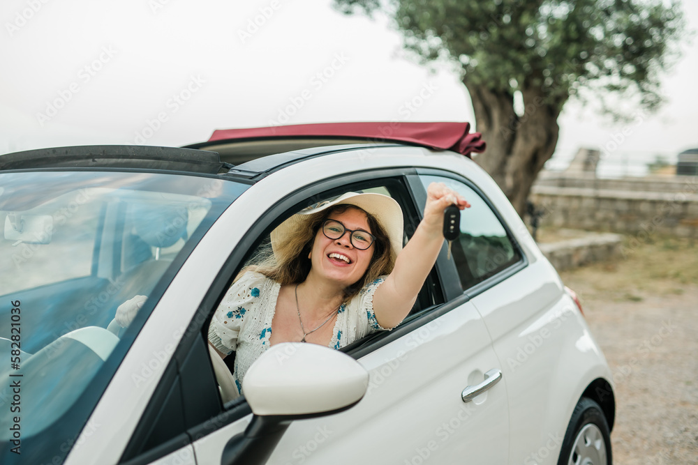 Car driver woman smiling showing new car keys and car. Female driving rented cabrio on summer vacation