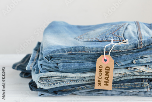 Stack of blue jeans and tag with inscription second hand on white background. Second hand clothing shop. Circular fashion, eco friendly sustainable shopping, thrift stores concept. photo