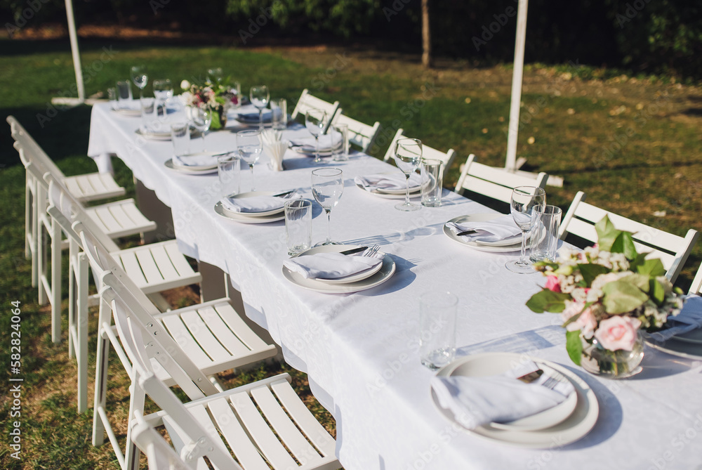 A large white long table with chairs, decorated with fresh flowers, with plates, glasses and forks, stands in a park with grass. Wedding decorations and details. Preparing for a wedding party.