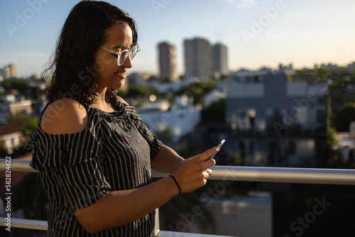 Close up of a smiling Latin girl standing on the balcony looking at mobile phone with a sunlight on her face