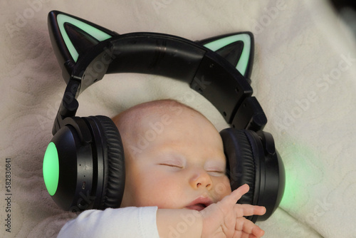 The infant sleeps in black earbuds in the shape of cat ears on a white bed.