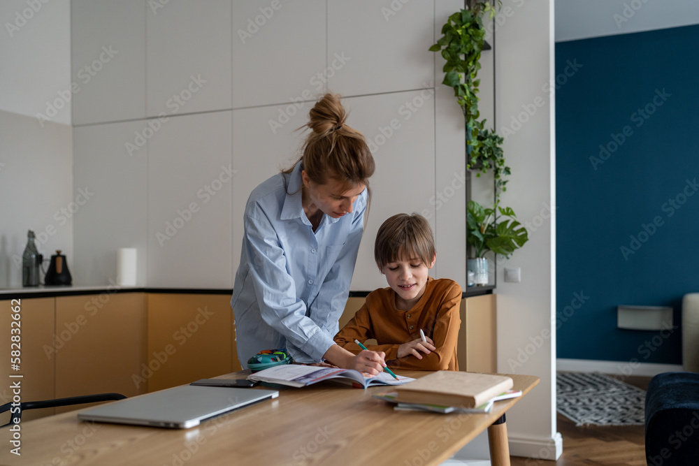 Young Caucasian mother helping child son with homework at home, sitting together at kitchen table. Happy schoolboy learning foreign language with private teacher tutor at home. Homeschooling concept Young Caucasian mother helping child son with homework at home, sitting together at kitchen table. Happy schoolboy learning foreign language with private teacher tutor at home. Homeschooling concept