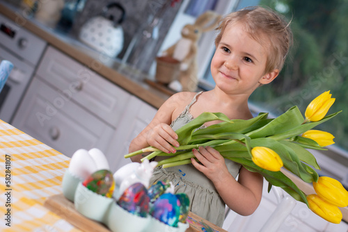 Little smilinggirl holding yellow flowers and decorated easter eggs on the table at home. Cute child and spring indoors. photo