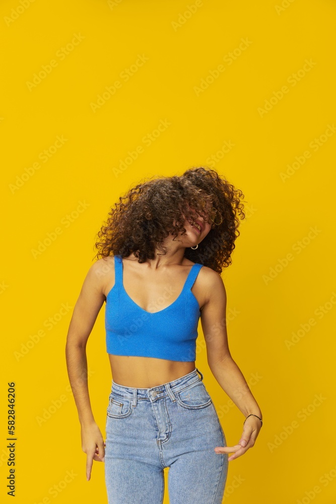 Woman with curly afro hair in a blue T-shirt on a yellow background dancing flying hair with sunglasses yellow, hand signs, look into the camera, smile with teeth and happiness, copy space Woman with curly afro hair in a blue T-shirt on a yellow background dancing flying hair with sunglasses yellow, hand signs, look into the camera, smile with teeth and happiness, copy space