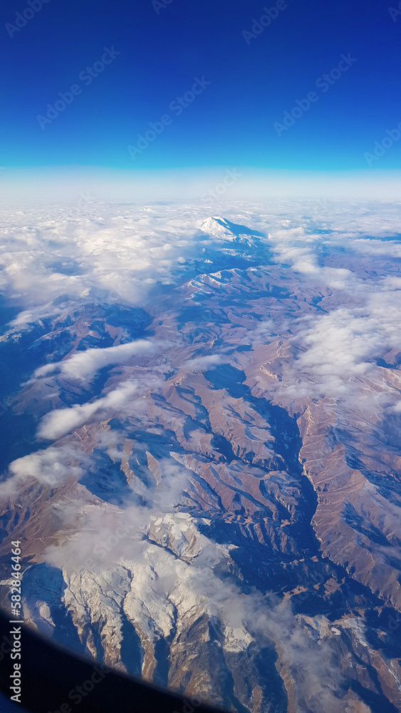 Fototapeta premium View from window of plane to blue sky and snowy mountain peaks in white clouds. High quality photo