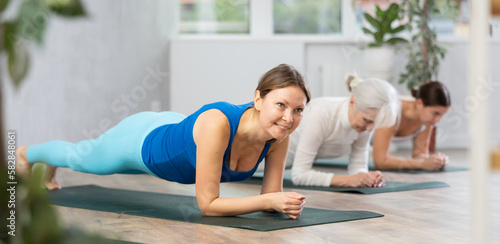 Diligent women practicing plank pose of yoga on black mat in light fitness room with house plants