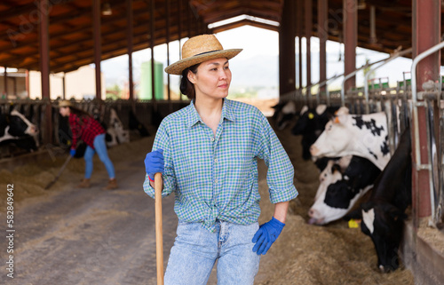 Cheerful asian woman farmer in straw hat who is standing at the farm against background of cows in stall