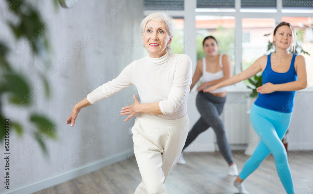 Cheerful sporty senior woman training movements of rhythmic latin zumba with group in fitness studio. Concept of active lifestyle of aged adults