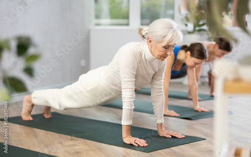 Concentrated elderly woman holding plank pose to strengthen body muscles during group yoga training in studio. Core exercises for older adults. Active lifestyle concept..