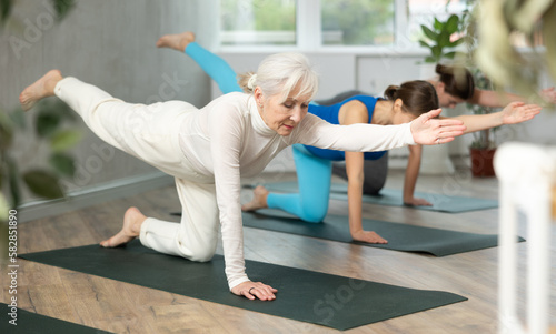 Concentrated fit senior woman maintaining active lifestyle exercising with group in yoga studio, standing in balancing asana Dandayamna Bharmanasana