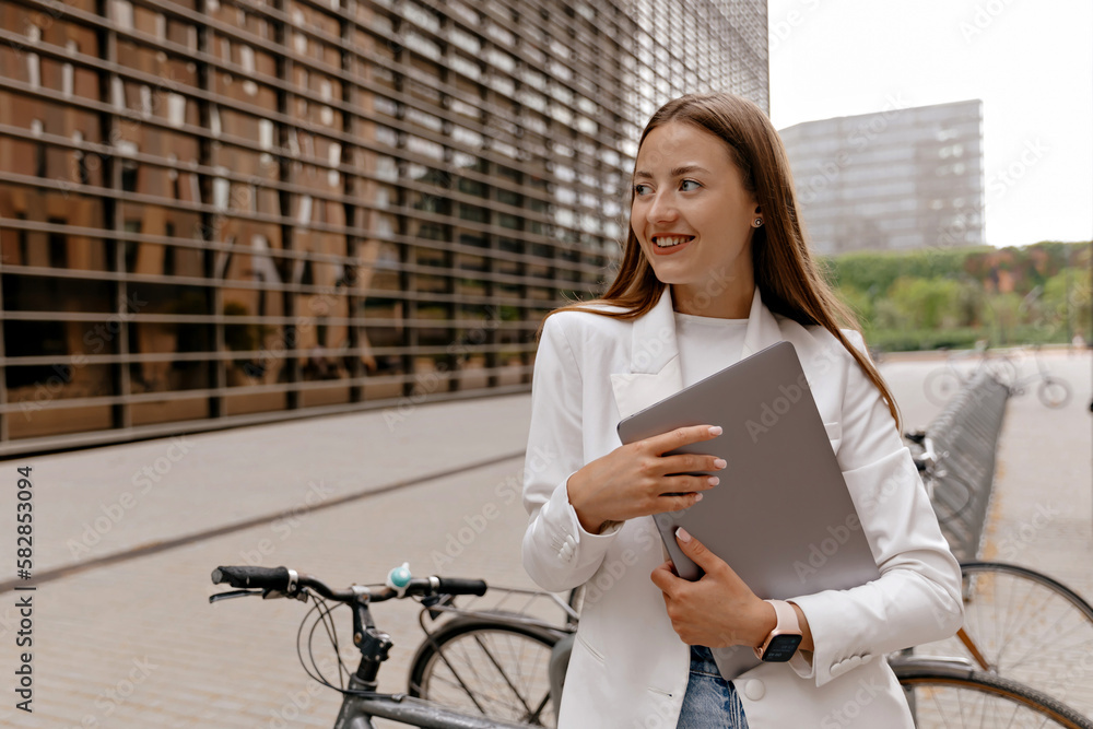 Cute lovely good-looking girl with happy smile wearing white jacket and jeans is holding laptop and posing to camera on background of modern buildings and city bikes