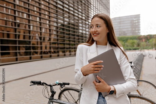 Obraz na płótnie Cute lovely good-looking girl with happy smile wearing white jacket and jeans is holding laptop and posing to camera on background of modern buildings and city bikes w salonie