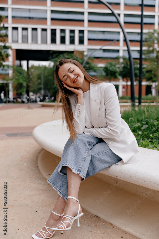 Fototapeta premium Full-length photo of beautiful relaxed girl wearing white jacket and jeans and heels is sitting outdoor on background of business district and resting with lovely smile and closed eyes. 