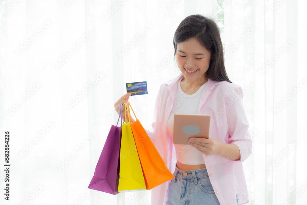 Young asian woman standing using digital tablet shopping online with credit card while holding paper bag, female paying with transaction financial, purchase and payment, business concept. Young asian woman standing using digital tablet shopping online with credit card while holding paper bag, female paying with transaction financial, purchase and payment, business concept.