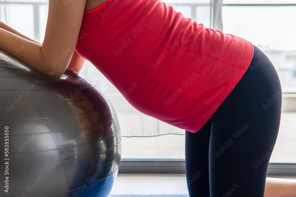 Pregnant woman in a vibrant sports shirt leaning on a physio or exercise ball and stretching against a window. Pregnancy, maternity, motherhood, sport, yoga, and health concept. Bright background.	
