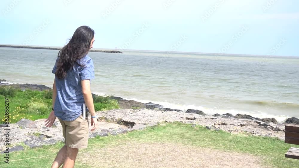 Pan shot of a young long haired latin man running towards a bench located on a river beach. Slow motion video. Pan shot of a young long haired latin man running towards a bench located on a river beach. Slow motion video.