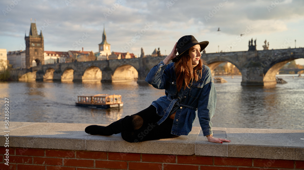 Stylish beautiful young woman wearing black hat sitting on Vltava river shore in Prague with Charles Bridge on background. Elegant retro lady fine art portrait.