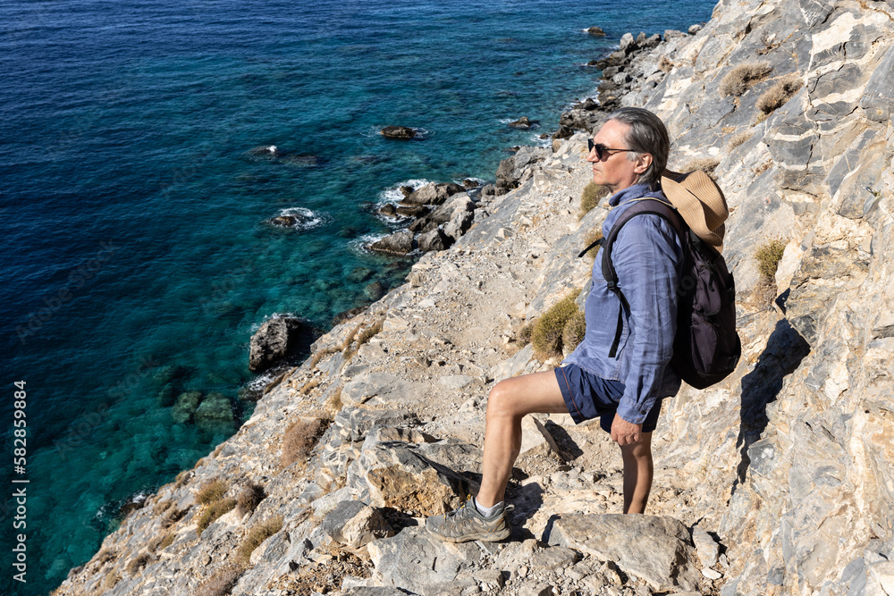 Grizzled long-haired man stands on a trail on a steep rocky shore, Greece, Crete