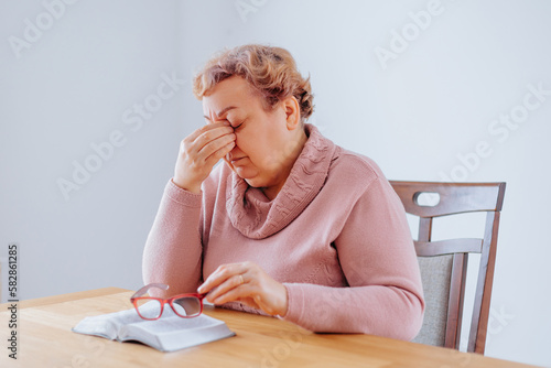 Depressed Senior Woman at Home Struggling with Eye Pain While Reading. A senior citizen sits at home with a book in her hand, her glasses perched on her nose as she struggles to read.