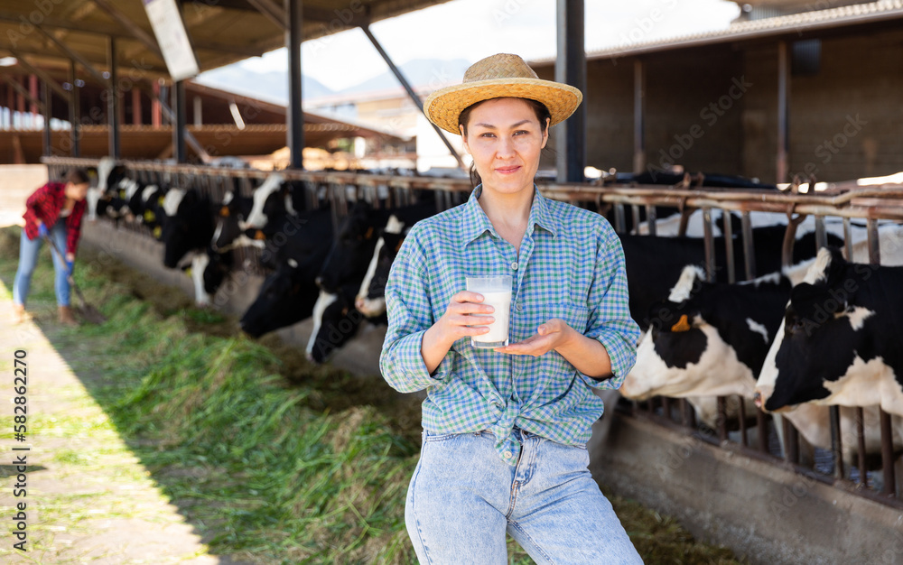 Portrait of a positive kazakh farmer woman standing in a cowshed, holding a glass of fresh milk in her hand