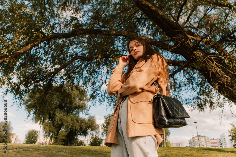 Caucasian young pretty stylish woman standing street pier near big huge tree dressed brown trench coat hold handbag smiling poses outside city, spring autumn season. Cute brunette hair lady
