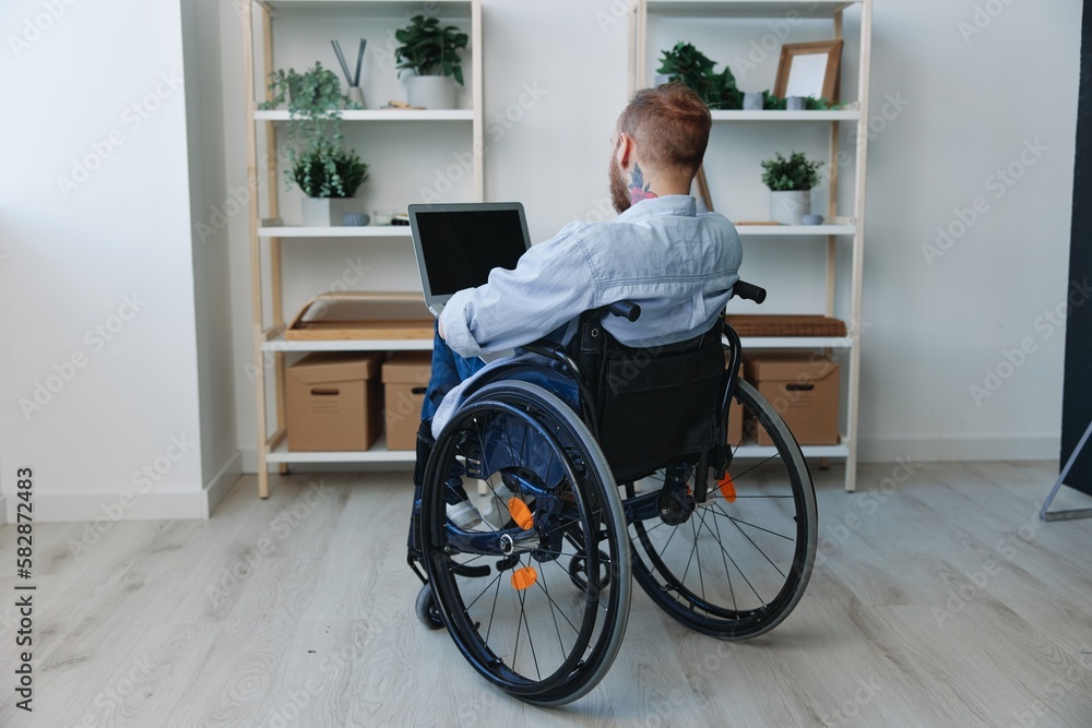 A man in a wheelchair freelancer works at a laptop at home, restriction of movement, view from the back, copy space, integration into society, health concept a person with disabilities, a real person A man in a wheelchair freelancer works at a laptop at home, restriction of movement, view from the back, copy space, integration into society, health concept a person with disabilities, a real person