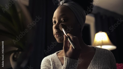 Black woman doing make up in dark room interior.African afro girl applying cosmetics on face while looking at mirror.Lady holding face powder beauty procedure in home studio