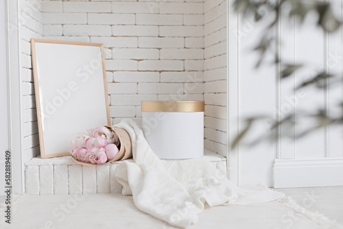 Spring and summer wedding concept scene, empty picture frame mockup on fireplace. Scandinavian interior style in white colour. Blooming peony bouquet.