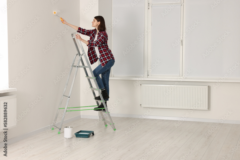 Young woman painting wall with roller on stepladder indoors. Room renovation Young woman painting wall with roller on stepladder indoors. Room renovation