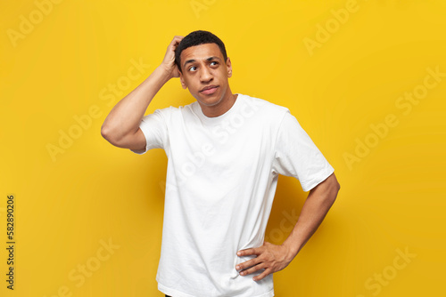 young puzzled african american guy in white t-shirt sketches his head with his hand on yellow isolated background